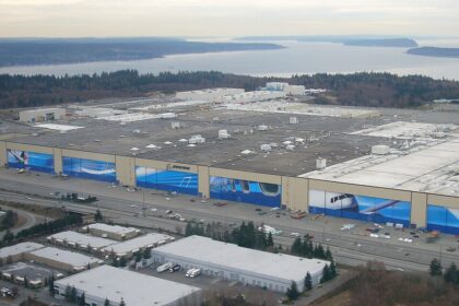 An aerial view of a massive Boeing manufacturing facility, characterised by its sprawling flat roof and enormous hangar doors adorned with large, blue aviation-themed murals and the Boeing logo. The massive complex is situated among smaller white-roofed industrial buildings, parking lots, and roads in the foreground, with a landscape of forested areas, a wide body of water, and distant landmasses visible in the background under a hazy sky.
