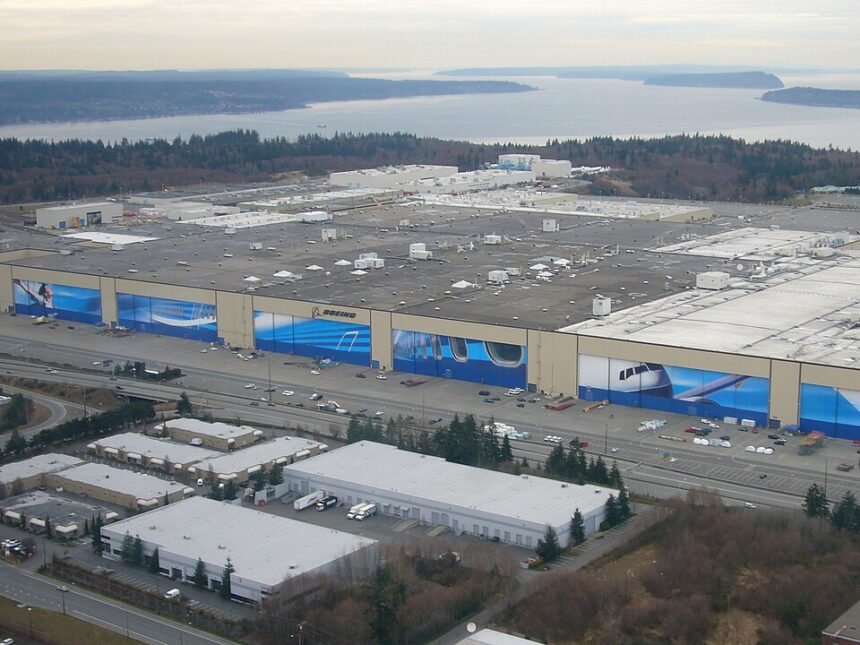 An aerial view of a massive Boeing manufacturing facility, characterised by its sprawling flat roof and enormous hangar doors adorned with large, blue aviation-themed murals and the Boeing logo. The massive complex is situated among smaller white-roofed industrial buildings, parking lots, and roads in the foreground, with a landscape of forested areas, a wide body of water, and distant landmasses visible in the background under a hazy sky.
