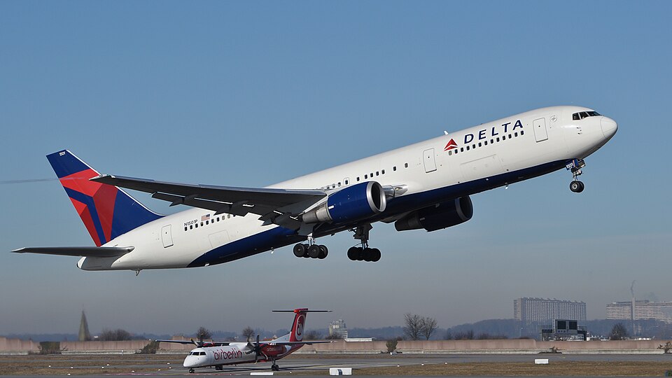 A Delta Air Lines Boeing 757 ascending steeply with landing gear down, contrasted against a clear blue sky, with a smaller Air Berlin aircraft visible on the runway in the background below.