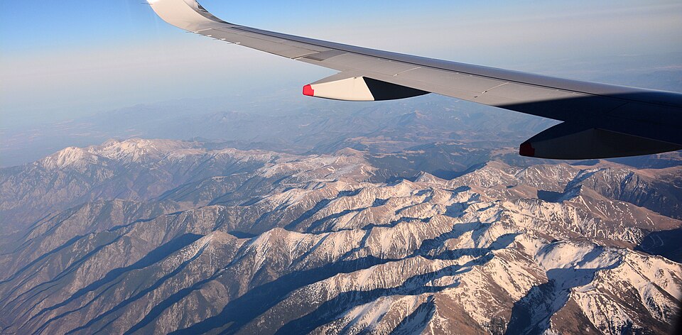 An aeroplane wing and engine visible through a window, high above a vast expanse of rugged, snow-capped mountains under a clear blue sky with a hazy horizon.