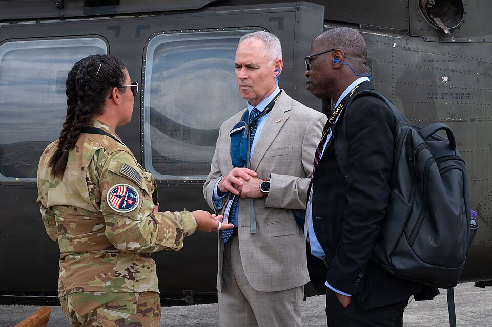 A U.S. military service member in camouflage speaks with two delegates wearing lanyards and ear protection in front of a military helicopter at the Farnborough International Airshow