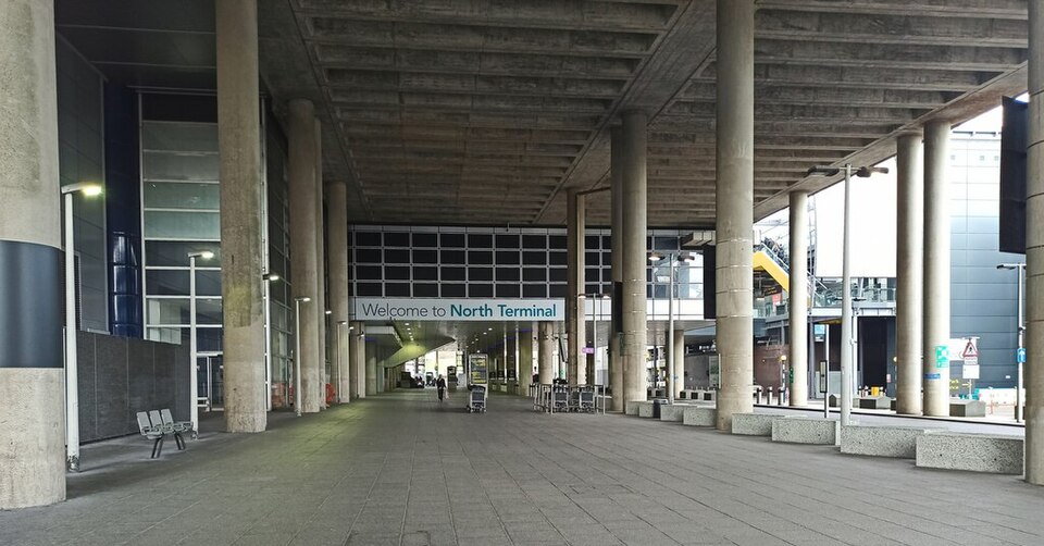 A ground-level, wide-angle shot of a spacious, covered pedestrian plaza outside the North Terminal at Gatwick Airport. A heavy, industrial-style brutalist concrete ceiling with a gridded pattern looms overhead, supported by a long colonnade of massive, smooth concrete pillars that stretch into the background.