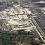 A high-resolution aerial view of London Gatwick Airport in West Sussex, England, showing the airport's single main runway running diagonally through the image. Visible are the North and South Terminals, extensive aircraft parking stands, taxiways, service roads, car parks filled with vehicles, and surrounding green countryside.