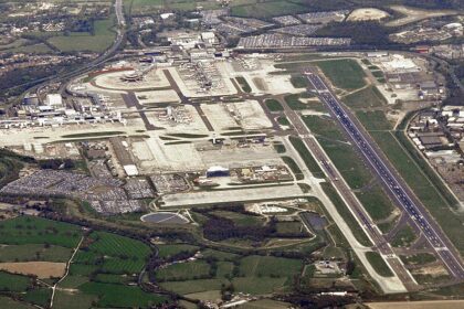 A high-resolution aerial view of London Gatwick Airport in West Sussex, England, showing the airport's single main runway running diagonally through the image. Visible are the North and South Terminals, extensive aircraft parking stands, taxiways, service roads, car parks filled with vehicles, and surrounding green countryside.