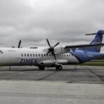 A detailed, side-angle view of a twin-engine Zimex Aviation ATR 72 regional airliner parked on a grey asphalt tarmac. The plane features a white forward fuselage with the blue text "ZIMEX" and a bright blue rear section and tail fin bearing the Swiss flag next to the registration "HB-ALM".
