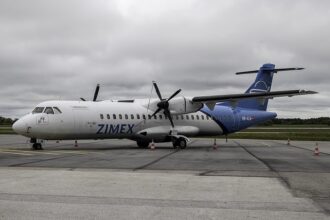 A detailed, side-angle view of a twin-engine Zimex Aviation ATR 72 regional airliner parked on a grey asphalt tarmac. The plane features a white forward fuselage with the blue text "ZIMEX" and a bright blue rear section and tail fin bearing the Swiss flag next to the registration "HB-ALM".