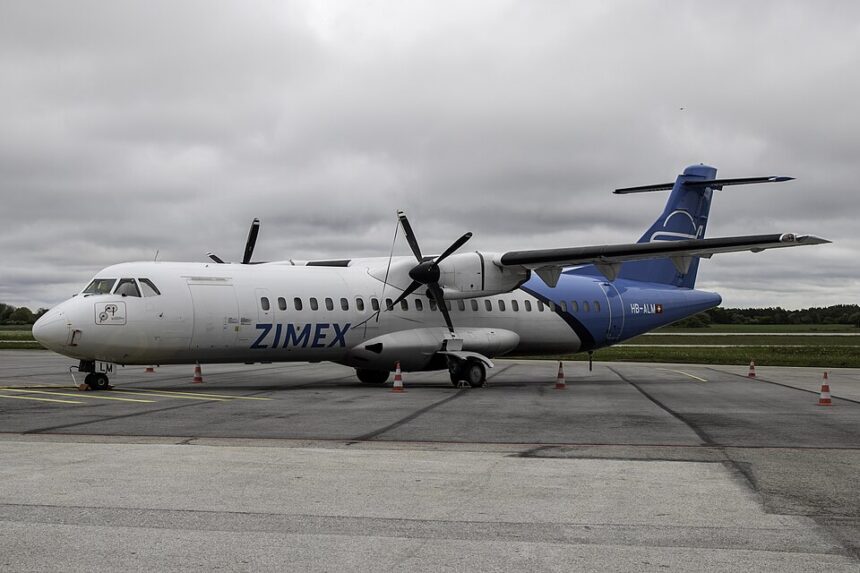 A detailed, side-angle view of a twin-engine Zimex Aviation ATR 72 regional airliner parked on a grey asphalt tarmac. The plane features a white forward fuselage with the blue text "ZIMEX" and a bright blue rear section and tail fin bearing the Swiss flag next to the registration "HB-ALM".