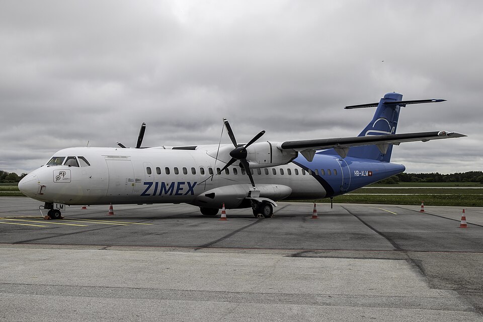 A detailed, side-angle view of a twin-engine Zimex Aviation ATR 72 regional airliner parked on a grey asphalt tarmac. The plane features a white forward fuselage with the blue text "ZIMEX" and a bright blue rear section and tail fin bearing the Swiss flag next to the registration "HB-ALM".