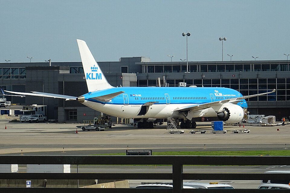 A KLM B787- parked outside of DUL's terminal A. The aircraft is half white and half blue, with its tail displaying the airline's logo in blue.