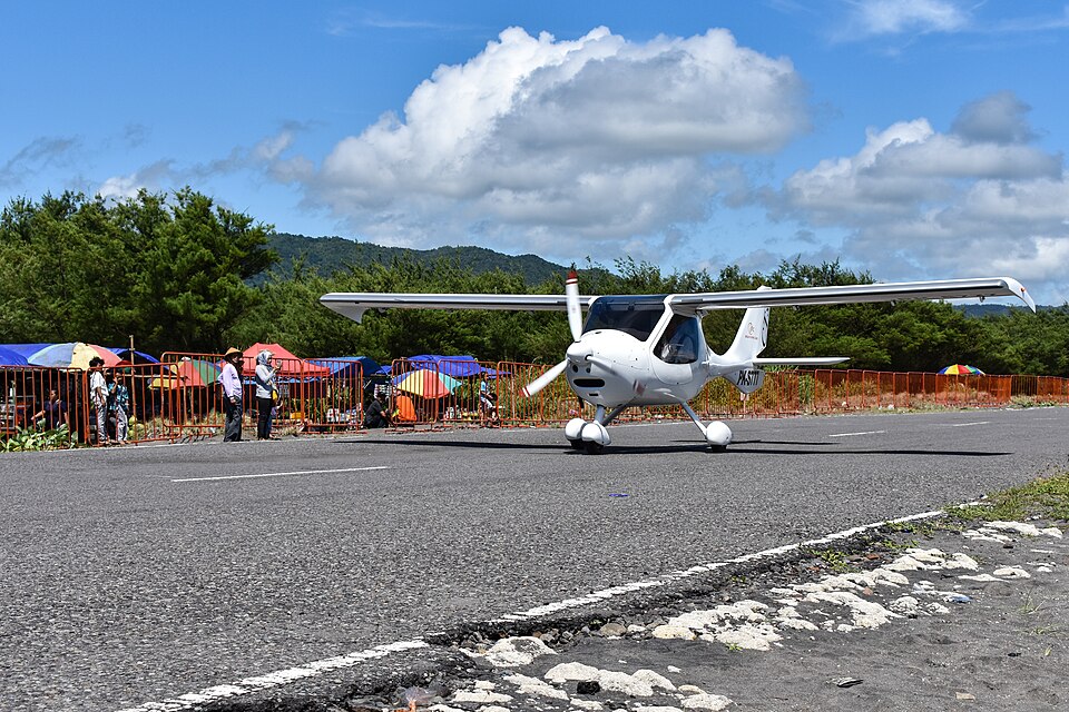 A small, white, single-engine high-wing light aircraft taxiing along an asphalt runway or paved road. To the left, a crowd of onlookers stands behind an orange metal crowd-control barricade, with several colorful sun umbrellas set up behind them. The scene is backed by lush green trees and forested hills under a bright blue sky with large, fluffy white clouds, while the rough, rocky edge of the pavement is visible in the bottom right foreground.