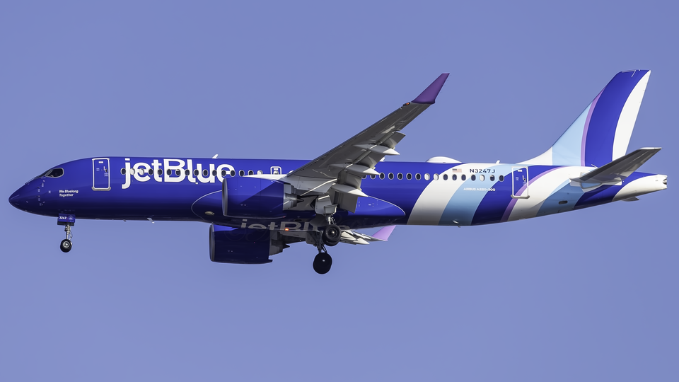 An JetBlue A220 flying under a blue sky. The aircraft's livery is dark blue, with its tail being streaks of light, dark blue and white.