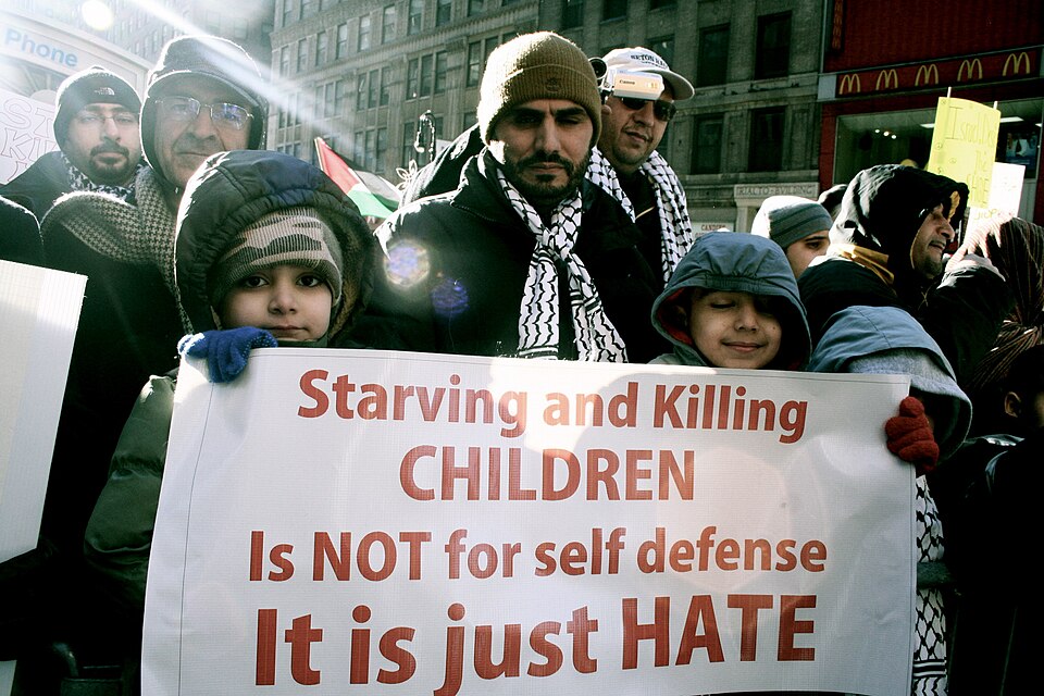 Image shows a large crowd at a Free Gaza protest demonstration in New York on January 3 2009. Two children are holding a white sign and looking at the camera with an adult between them. The white sign has red writing and reads 'Starving and Killing CHILDREN Is NOT for self defense It is just HATE'