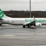 A Transavia Boeing 737-800 (registration N893SM) parked on a wet airport tarmac, featuring the airline's white livery with a large green "t" logo on the tail and "transavia" titles in green lettering along the fuselage, with another aircraft partially visible in the background.