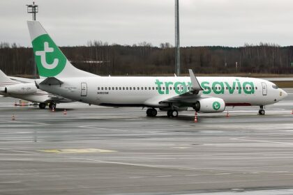 A Transavia Boeing 737-800 (registration N893SM) parked on a wet airport tarmac, featuring the airline's white livery with a large green "t" logo on the tail and "transavia" titles in green lettering along the fuselage, with another aircraft partially visible in the background.