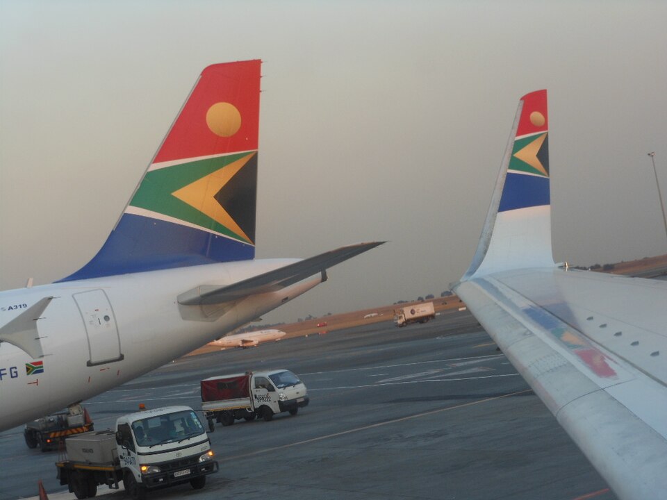 A detailed shot capturing the distinctive branding of South African Airways on the tarmac. On the left, the rear fuselage and vertical stabilizer of an Airbus A319 show the colorful tail livery adapted from the South African national flag, featuring red, blue, green, yellow, and black. 