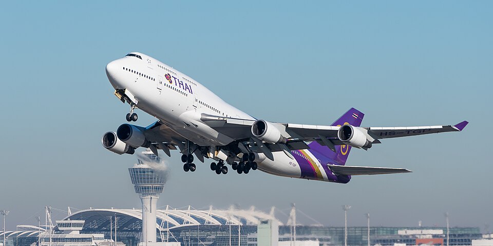 A Thai Airways Boeing 747-400 taking off, climbing steeply into a clear blue sky with its landing gear extended, and an airport control tower is visible in the background below.