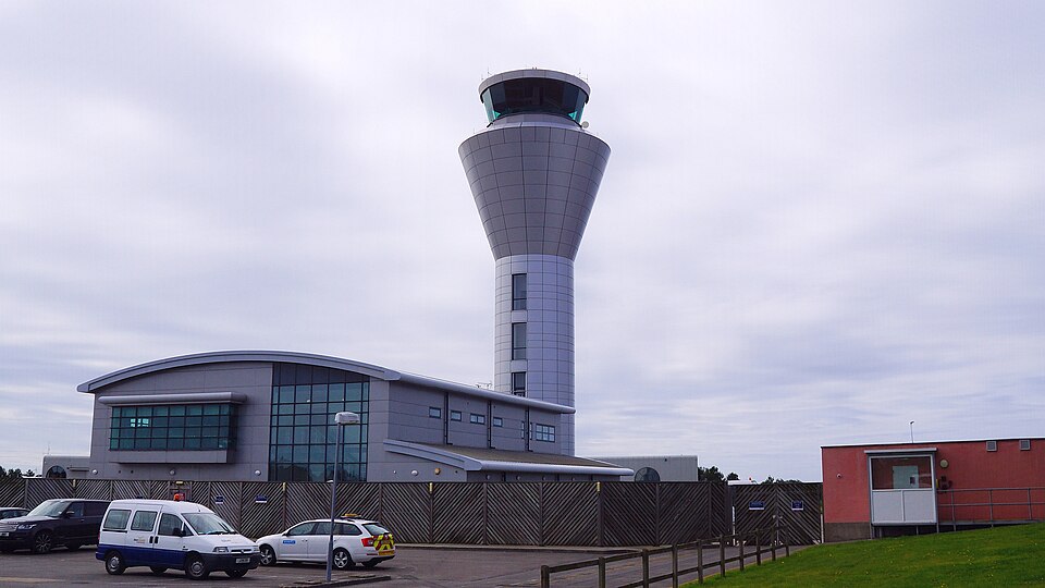 Tower building at Jersey Airport, home of the Jersey-Heathrow route. The sky is grey and there are two cars parked in front of the tower