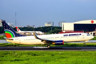 A U.S.-Bangla Airlines Boeing 737 (registration S2-AJB) landing on a runway, with white smoke billowing from its rear tires upon touchdown against a backdrop of green grass and airport buildings.