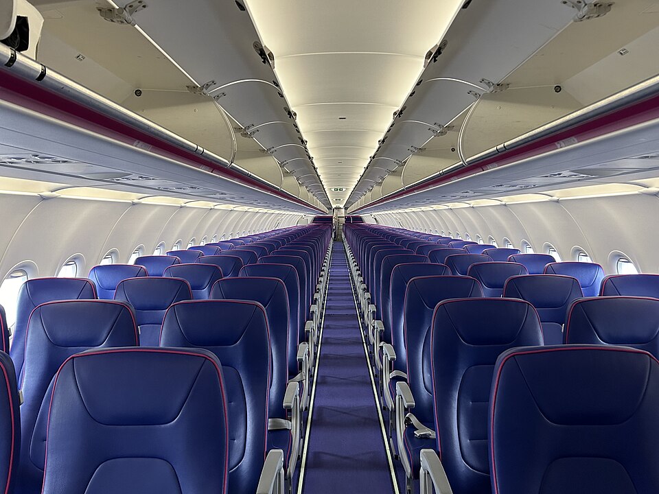 The interior of a Wizz Air Airbus aircraft is shown with neatly arranged blue leather seats, a central aisle, and overhead luggage compartments. The bright, modern cabin reflects the airline’s low-cost, high-capacity configuration used across its European network.