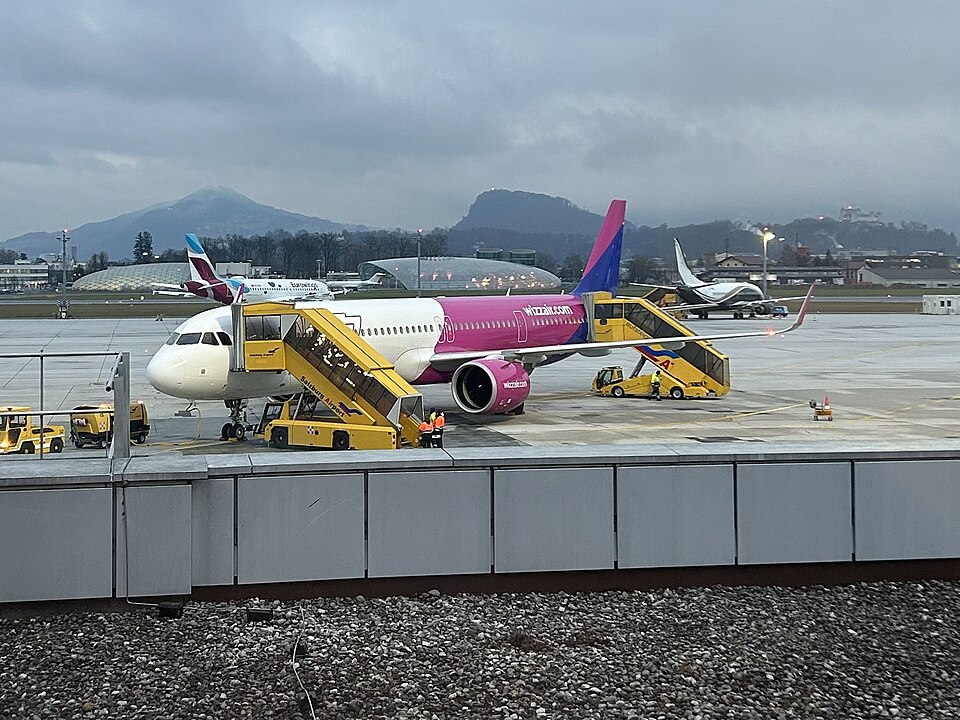 A Wizz Air Airbus A321 stands at a gate at Salzburg Airport during ground handling operations. Passenger boarding stairs and airport service vehicles surround the aircraft, while mist-covered mountains and cloudy skies.