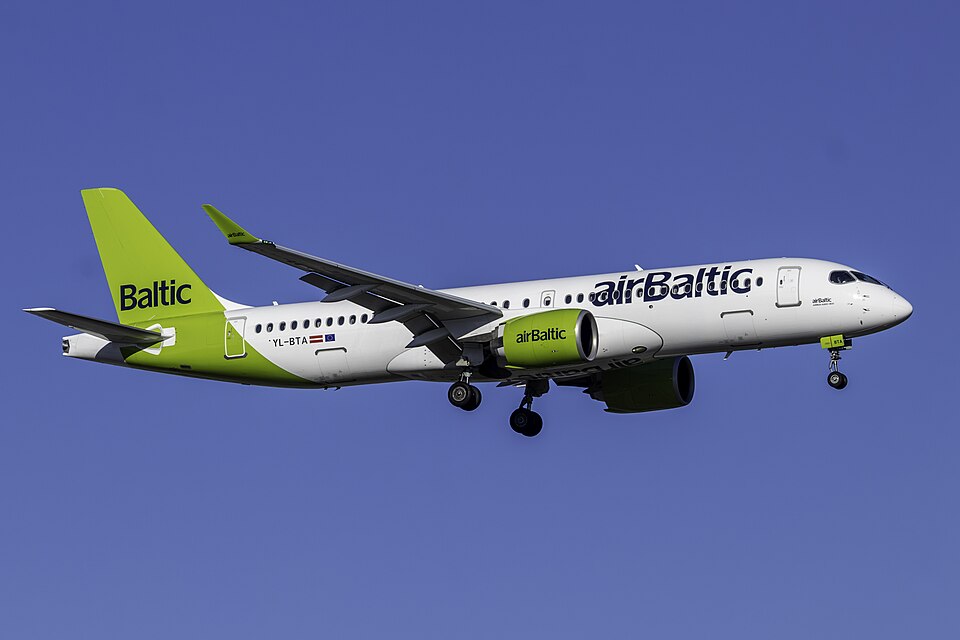 An airBaltic Airbus A220-300 passenger airliner (registration YL-BTA) with landing gear deployed descends against a clear blue sky.
