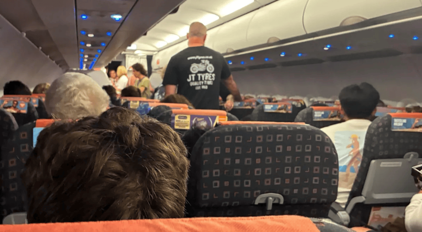 Passengers seated inside an EasyJet aircraft. A few passengers are standing in the aisle beneath overhead bins and blue cabin lights.