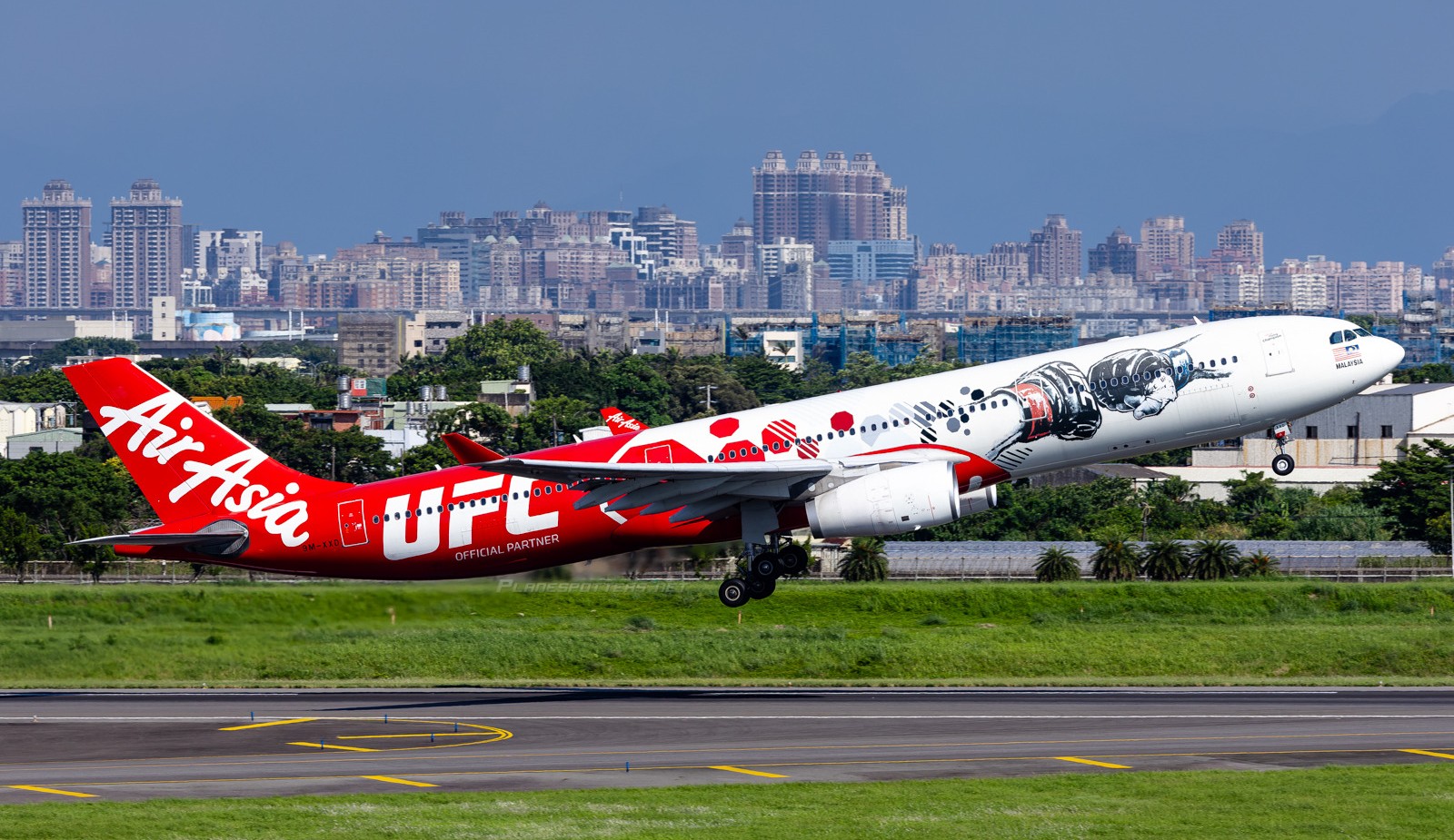 AirAsia X Airbus A330-343, registration 9M-XXD, in UFC partnership livery, climbing after takeoff with a city skyline in the background.