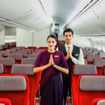 Inside of an Air India aircraft. Red seats with a headrest. Two crew members in the middle of the frame greeting the camera with their hands together.