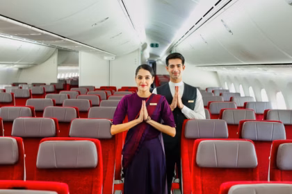 Inside of an Air India aircraft. Red seats with a headrest. Two crew members in the middle of the frame greeting the camera with their hands together.