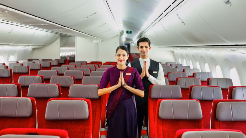 Inside of an Air India aircraft. Red seats with a headrest. Two crew members in the middle of the frame greeting the camera with their hands together.