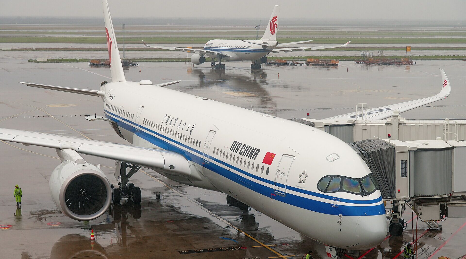 An Air China Airbus A350-900 parked up at the airport. Rain on the floor reflecting the aircraft. Another Air China aircraft visible in the background heading away from the camera.