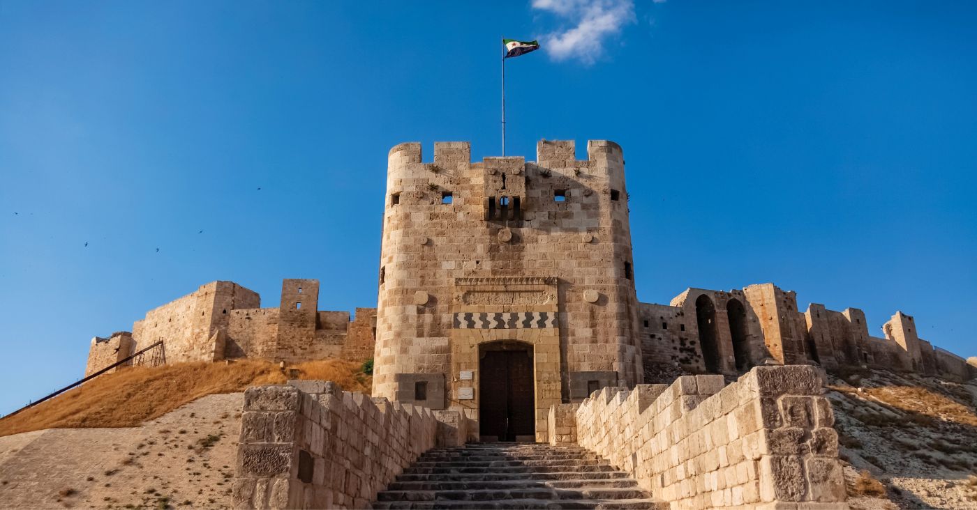 A stone-built Aleppo citadel with stairs at the bottom and a Syrian flag at the top with a clear sky background.