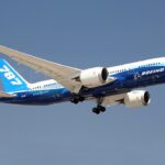 A Boeing 787 is pictured in flight, tail on the left side and nose on the right. The underbelly of the aircraft is cobalt blue with white 'BOEING' branding along it, and the tail is a light blue with white '787' branding. The rest of the jet is white. It is pictured at a slight upwards angle. The background is a clear blue sky.
