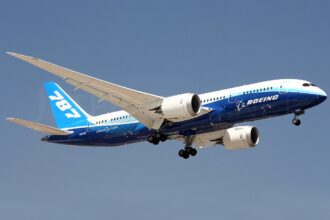 A Boeing 787 is pictured in flight, tail on the left side and nose on the right. The underbelly of the aircraft is cobalt blue with white 'BOEING' branding along it, and the tail is a light blue with white '787' branding. The rest of the jet is white. It is pictured at a slight upwards angle. The background is a clear blue sky.