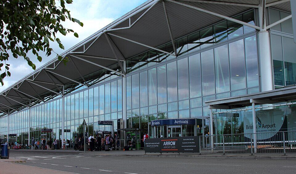 Exterior of Bristol Airport. Floor to ceiling windows shown the whole way across. Sign outside saying "Arrivals" and passengers seen outside of the building with luggage.