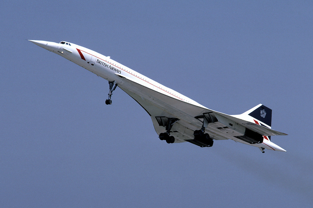 A British Airways Concorde pictured in flight during 1986. The aircraft is pictured ascending, pointed nose in the upper left of the image and the tail on the lower right. A red stripe runs across the middle of the entire body of the plane, and underneath the strict toward the front is dak blue branding which reads 'BRITISH AIRWAYS'. The tail also has navy blue, red, and white branding on the tail. The background is a clear blue sky.