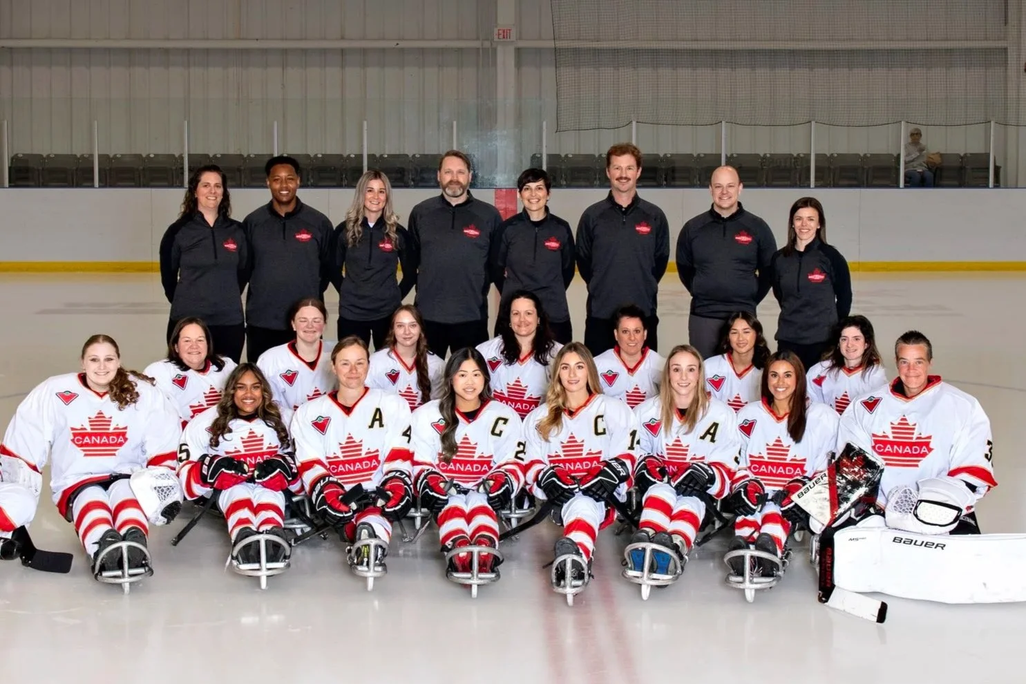 the women's para hockey team with players wearing white and red in the front and coaches stood behind wearing black. on the ice rink wearing all the hockey gear