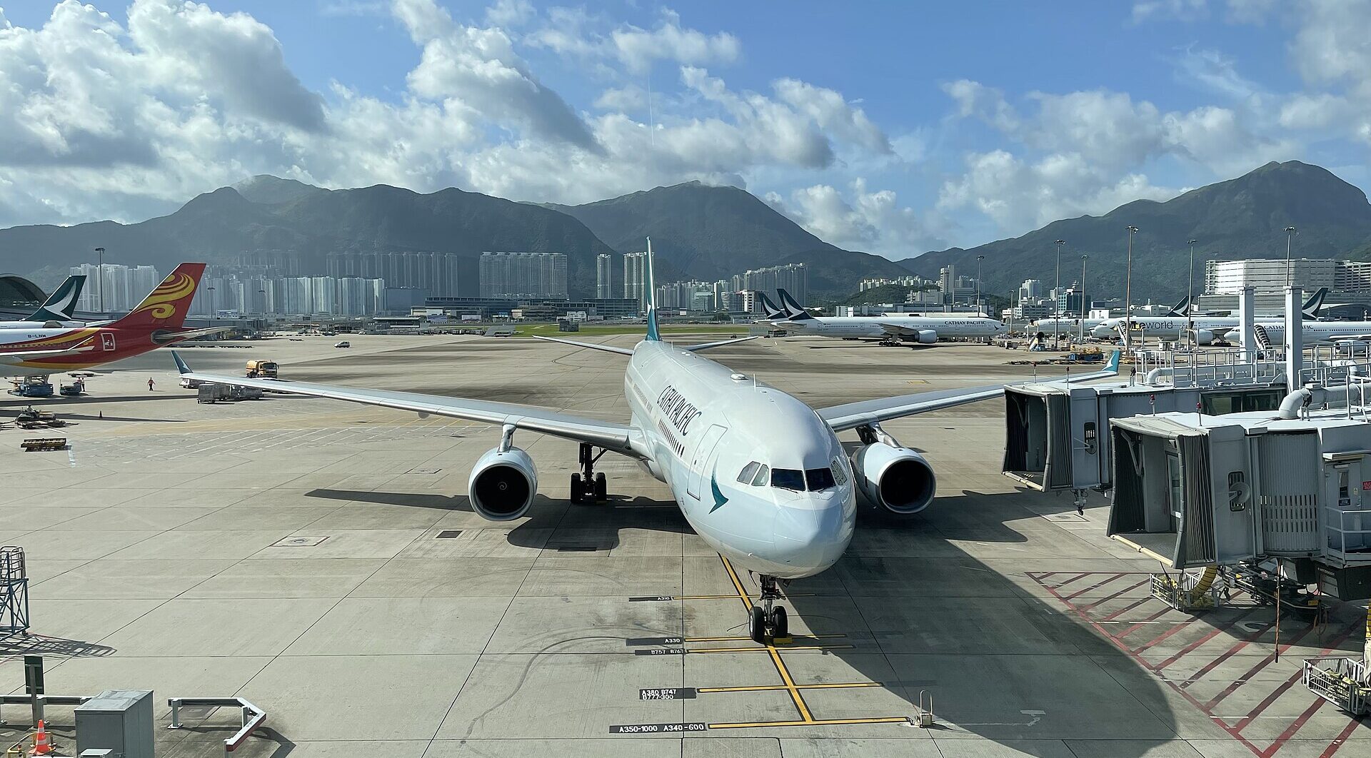 A Cathay Pacific plane in the apron at Hong Kong International Airport, facing toward the camera. Mountains seen in the background. Blue sky with clouds also seen across the top.