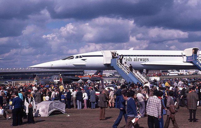 Image shows a British Airways Concorde aircraft grounded at the 1976 Farnborough Airshow on a cloudy day, with crowds of people underneath and people boarding the open doors