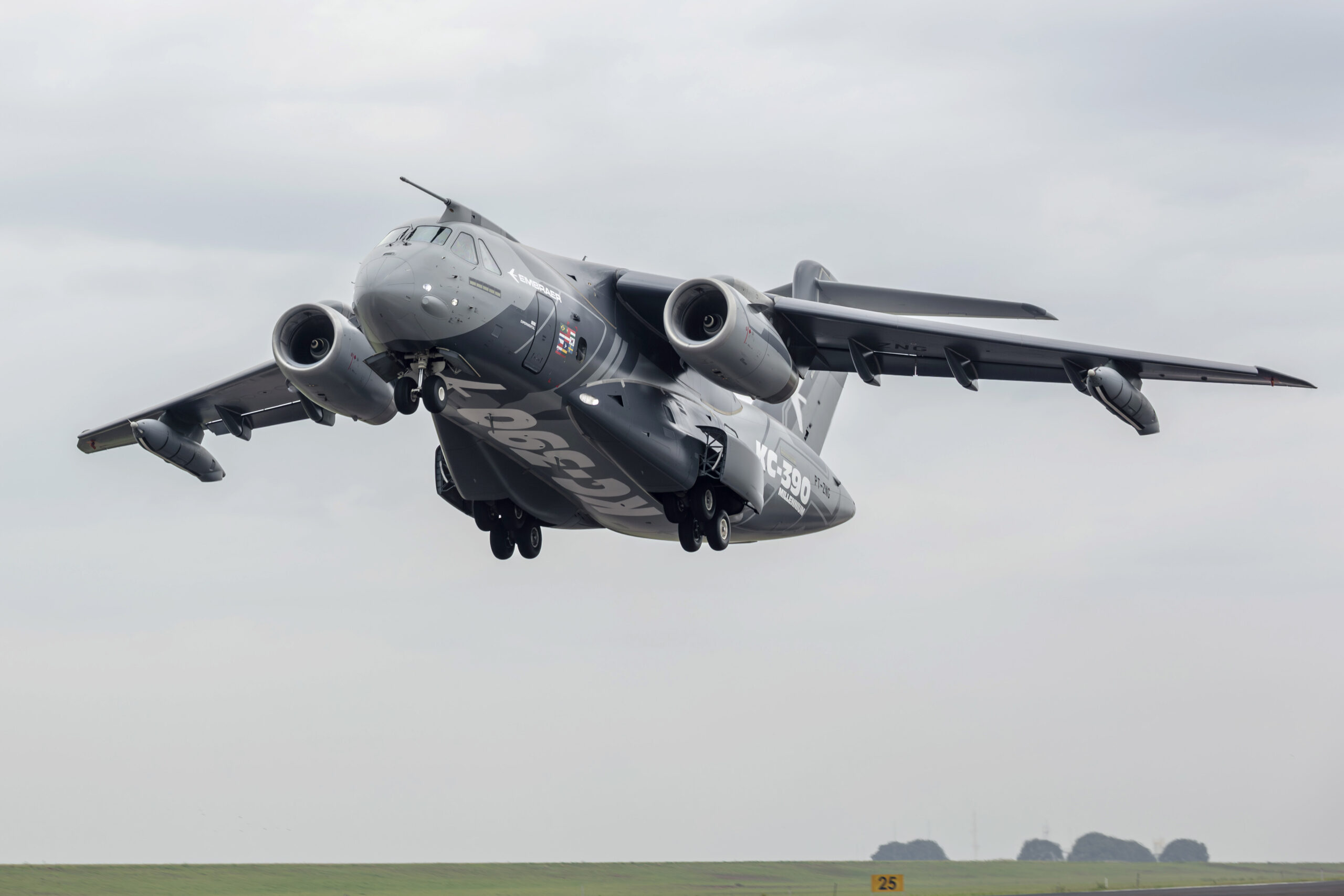 KC-390 Millennium grey aircraft flying above an airfield branded with its name and number against a grey sky