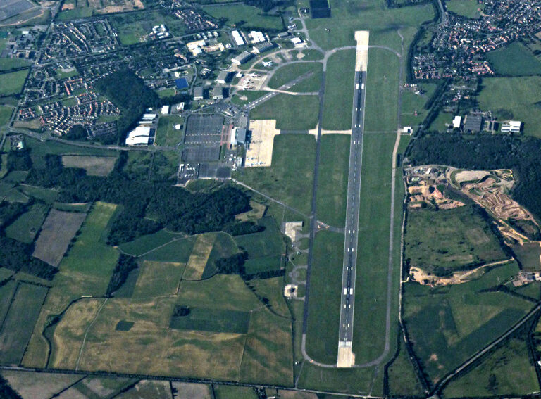 An aeiral photograph of Doncaster Sheffield Airport (DSA), taken in 2020. A grey tarmac runway can be seen centre right, surrounded by green fields and some residential looking areas a short distance away to the right. In the upper centre left of the image, the airport and its various buildings and facilities can be seen. 
