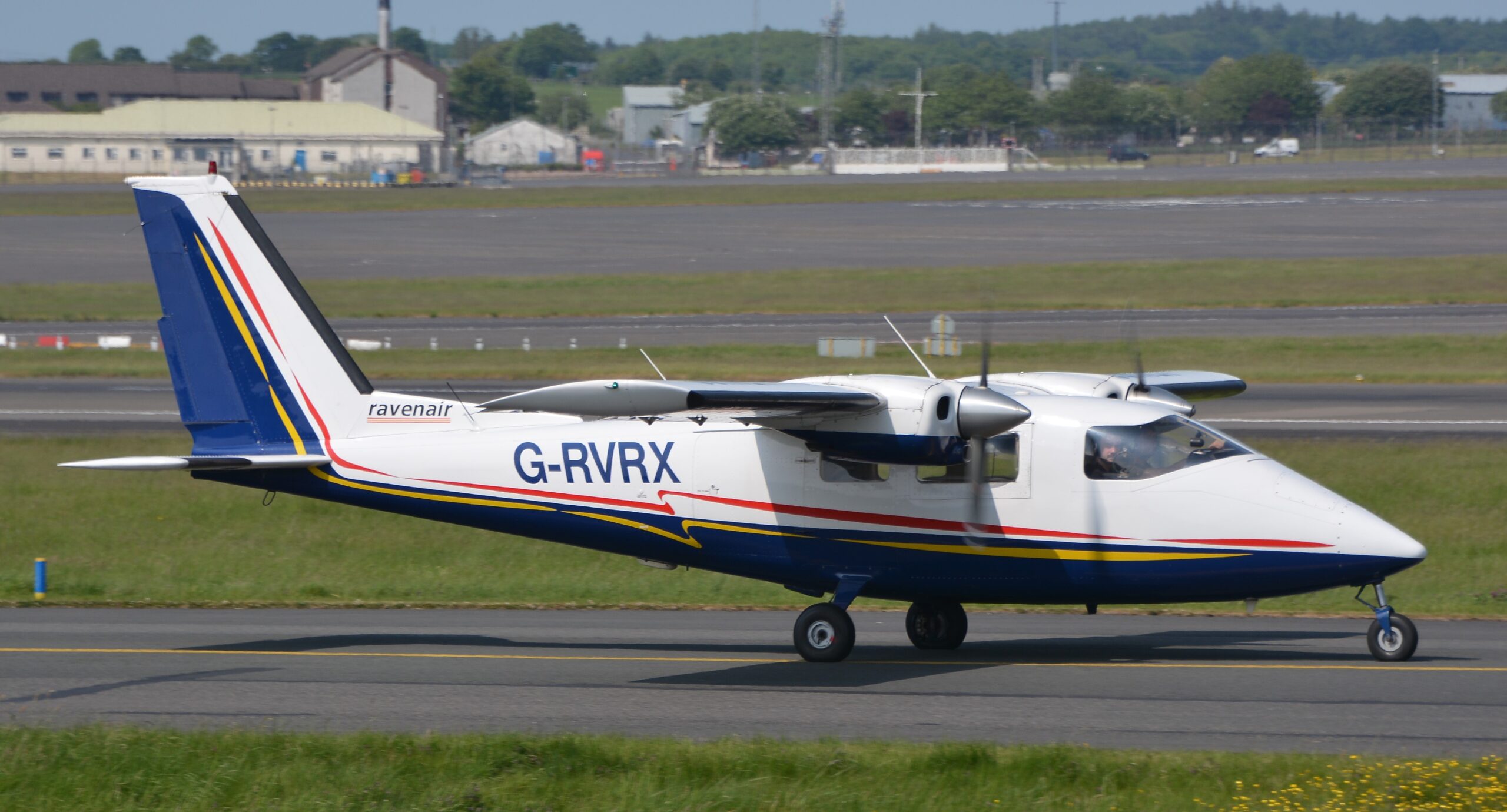 Side view of a Ravenair Partenavia P.68 twin‑engine light aircraft taxiing on an airport runway. The aircraft is painted white with dark blue, red and yellow stripes along the fuselage and tail, and carries the registration G‑RVRX and the Ravenair name near the rear. Both propellers appear blurred from rotation, while the nose and main landing gear remain on the ground. In the background, grassy areas, taxiways, airport buildings and distant trees stretch beneath a clear daylight sky.