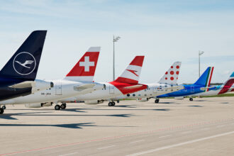 A set of jet tails displaying the logos of some of the Lufthansa Group’s subsidiary airlines. The nets are sat on an airport runway on a cloudy day.