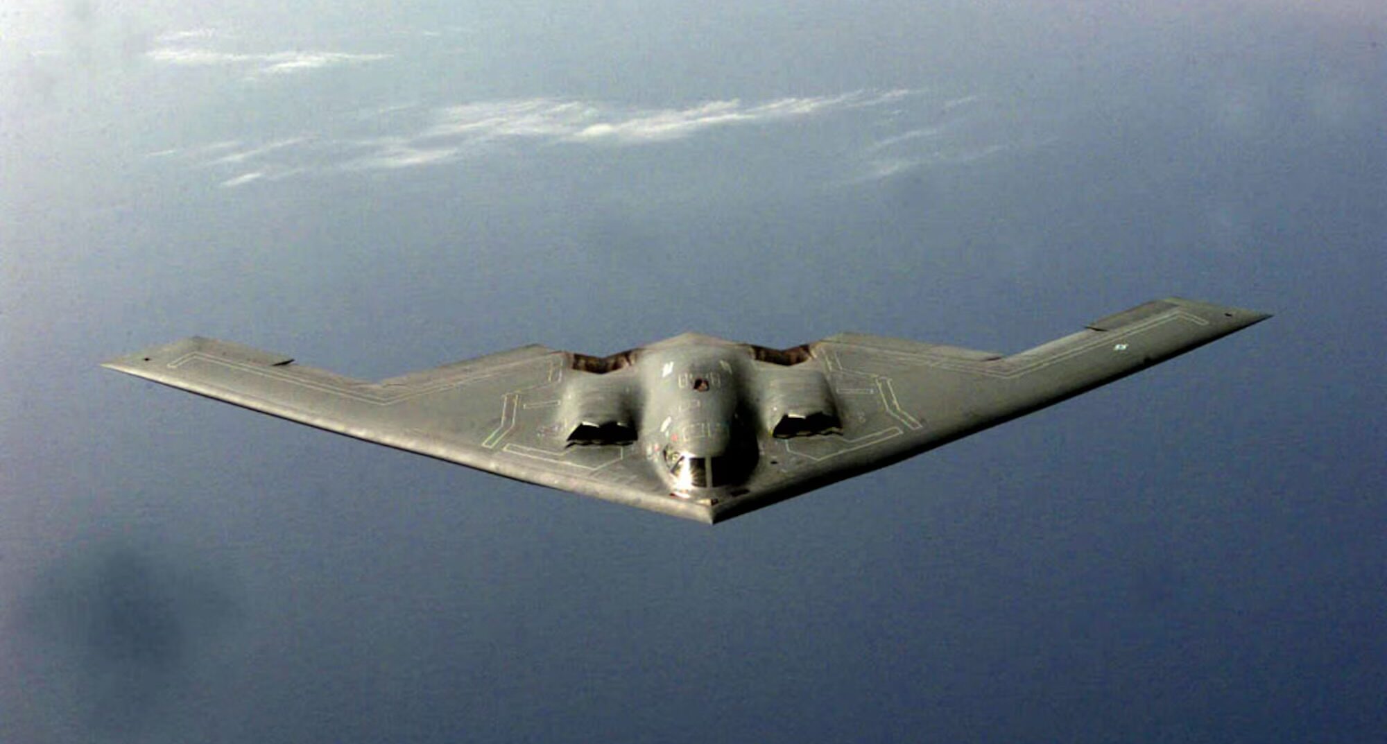 A dark grey Northrop B-2 Spirit flying over a dark blue ocean, with some white clouds behind it.