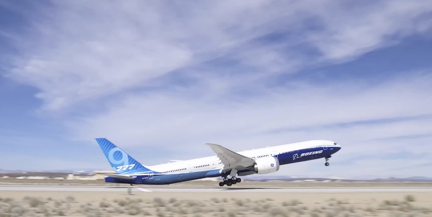 A blue and white Boeing 777X taking off from a runway in front of a cloudy sky.