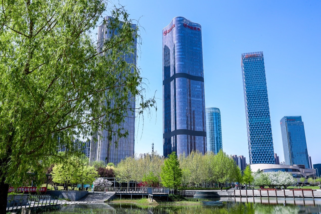 The Chaoyang district skyline photographed from the ground in front of a clear blue sky, with a lake in the foreground.