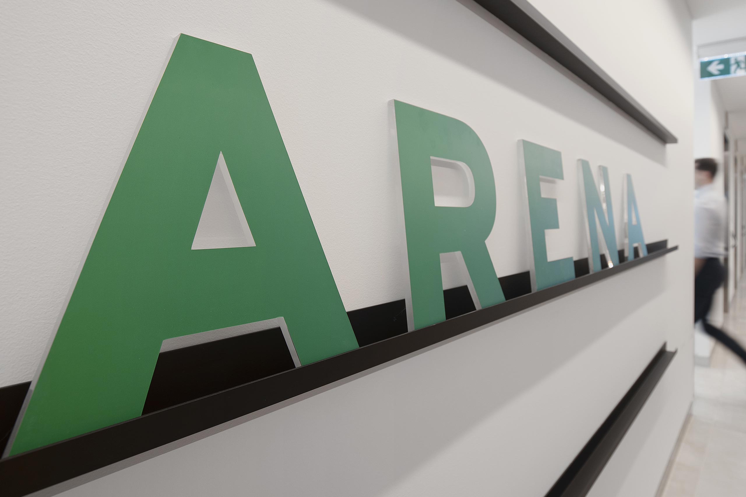 The word ARENA is spelt out in green capital letters along a white wall, each letter is sat on a black shelf.