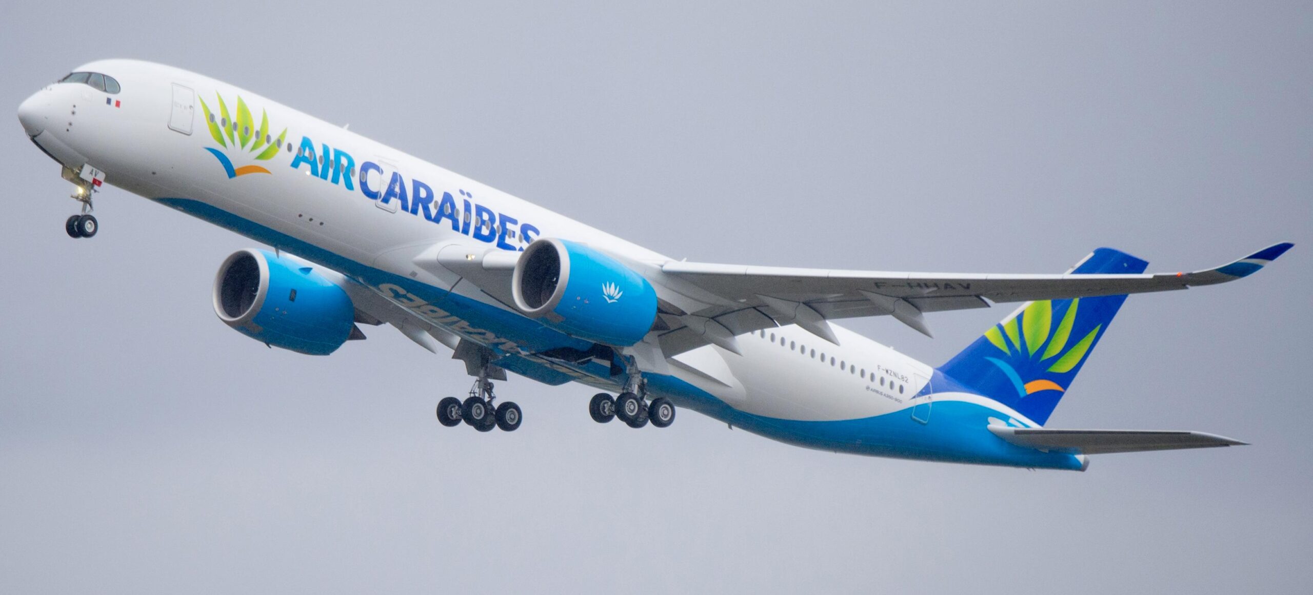 A blue and white Air Caraibes jet flying upwards in front of a grey sky.