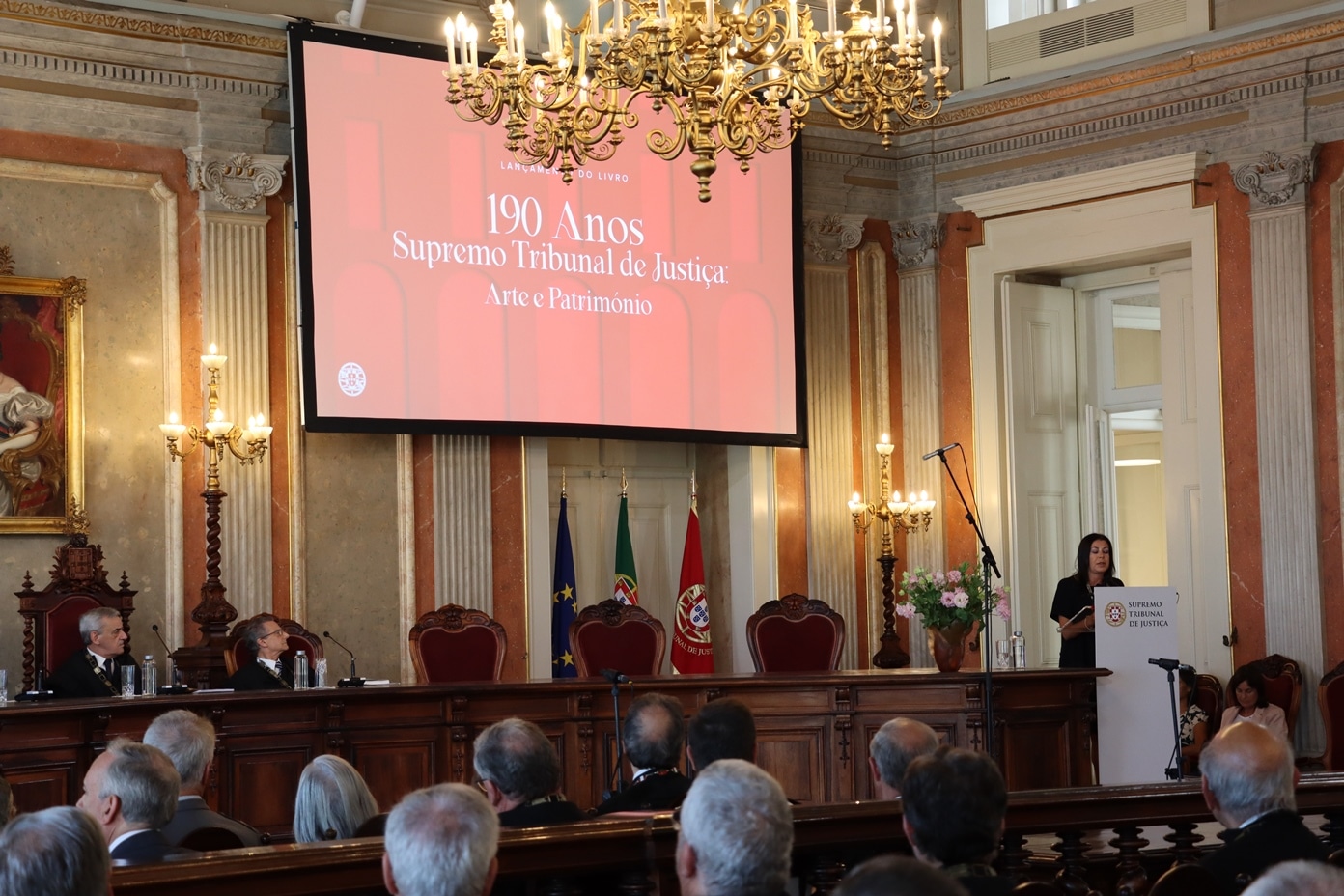 The Supremo Tribunal de Justicia de Portugal celebrating its 190th anniversary in 2023. A group of people are seen gathered in front of a red screen hanging from an ornately patterned ceiling.