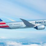 An red, white, and blue American Airlines jet flying in front of a cloudy sky.